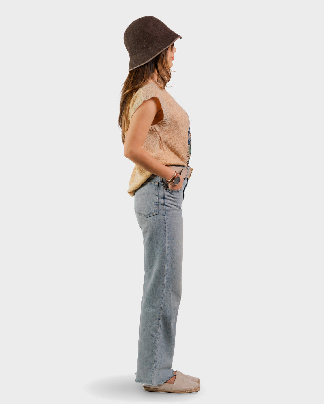 A woman shown in profile wearing a dark brown bucket hat, a beige sleeveless top, light-wash blue jeans, and beige slip-on shoes against a plain light gray studio background.