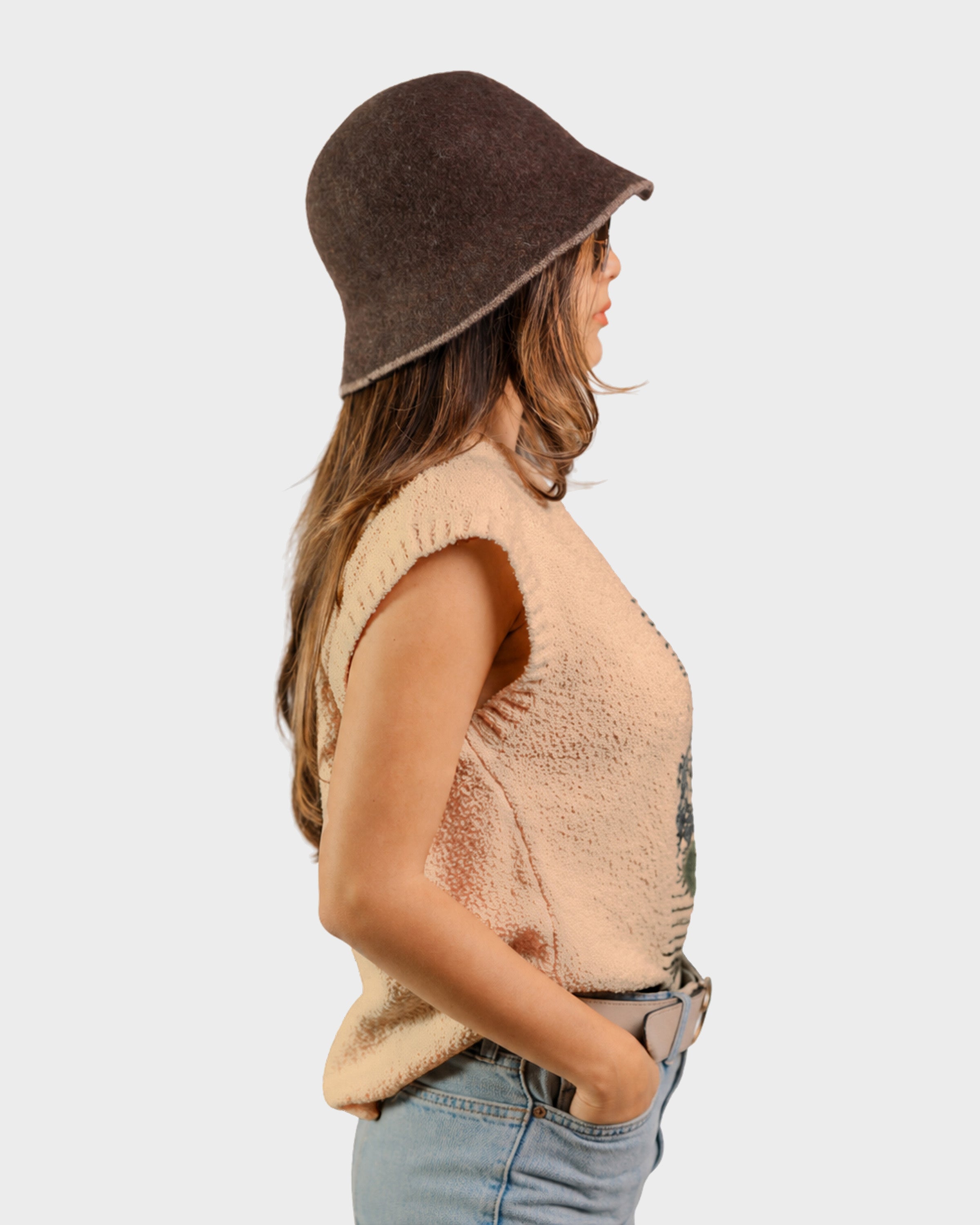 A woman shown in side profile wearing a dark brown felt bucket hat, a beige textured sleeveless top, and light blue jeans against a plain light gray background.