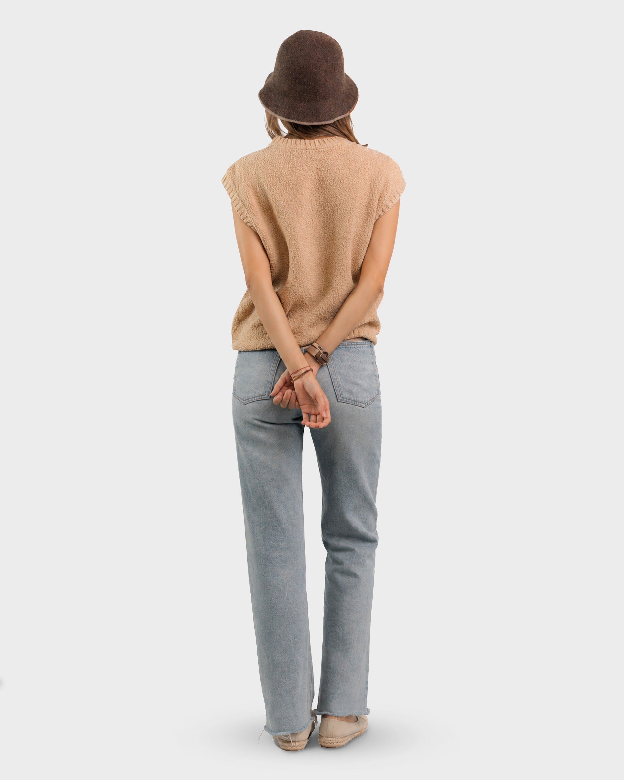 A woman shown from behind wearing a brown bucket hat, a beige sleeveless knit sweater vest, light blue jeans, and beige shoes, standing against a light gray studio background.