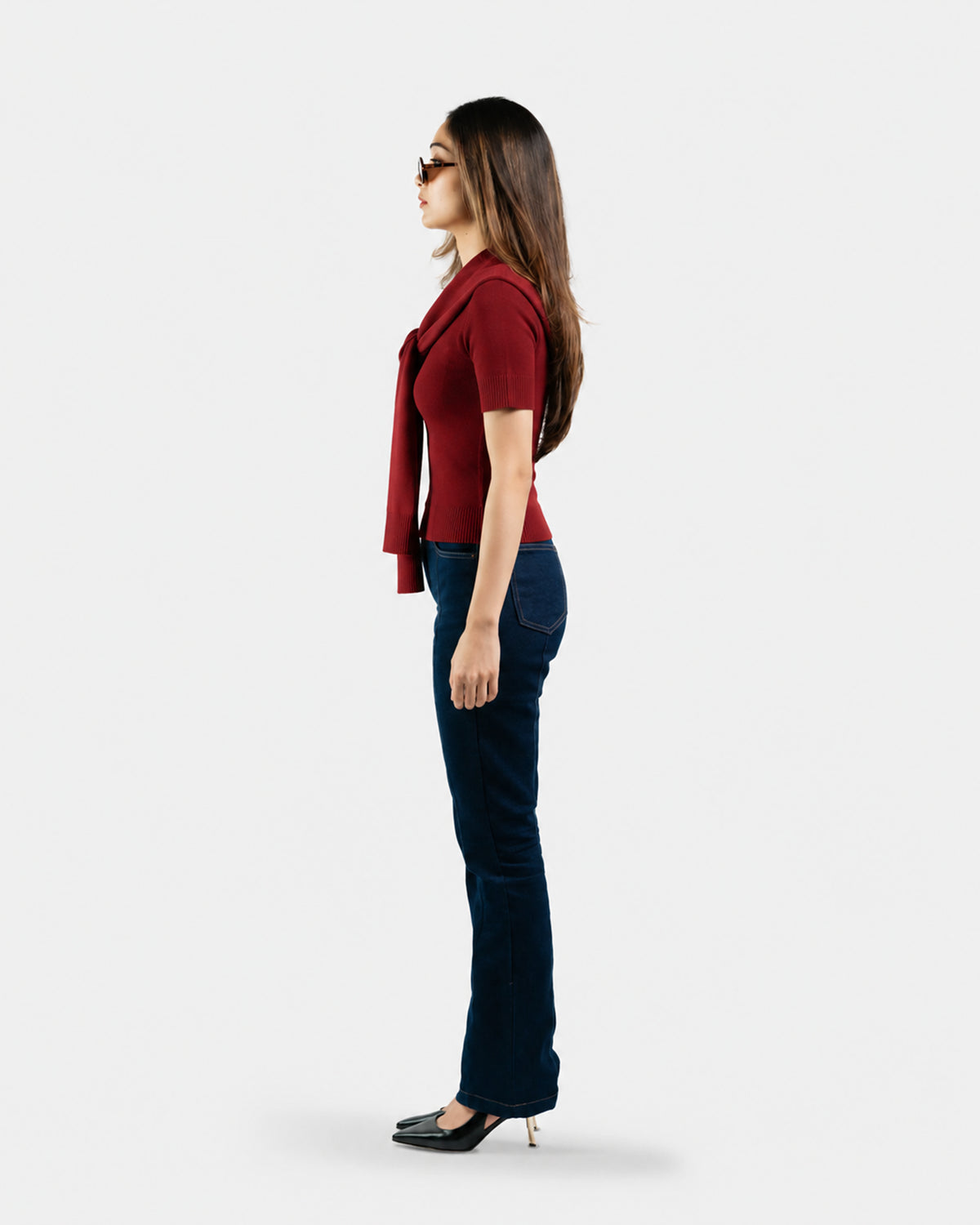 A woman shown in profile wearing a short-sleeve red knit top with a draped collar, dark blue jeans, and black pointed-toe heels against a plain light gray studio background.