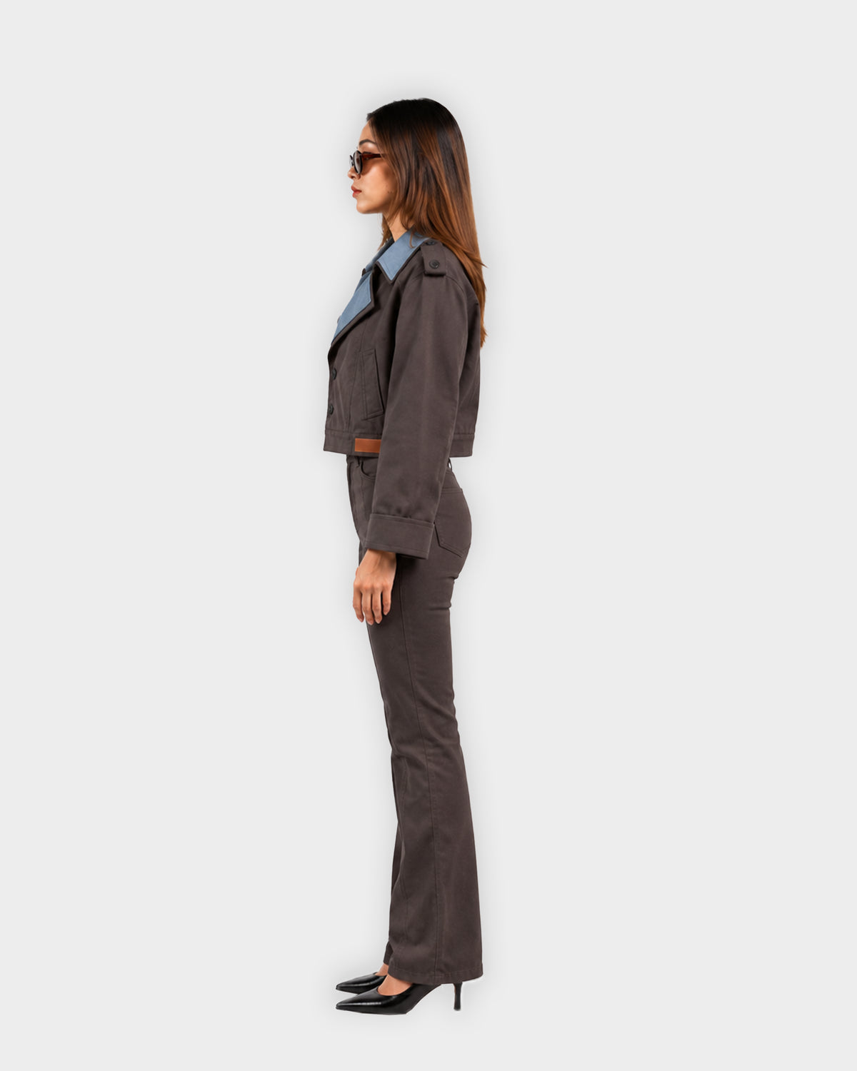 A woman stands in profile wearing a dark brown tailored suit with a cropped jacket, matching trousers, black pointed heels, and sunglasses against a plain light gray studio background.