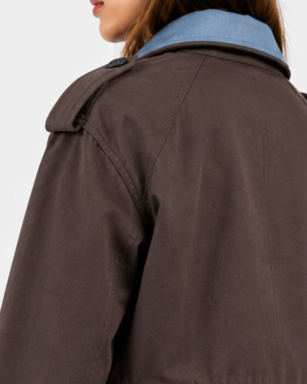 Close-up of a person wearing a dark brown trench coat over a light blue collared shirt, shown from the back against a plain light background.