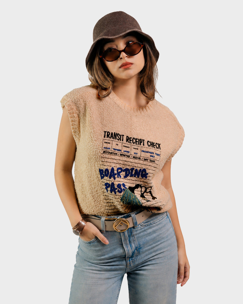 A woman wearing a beige sleeveless knit top with a vintage transit ticket graphic, styled with light blue jeans, a brown bucket hat, and sunglasses against a light gray background.
