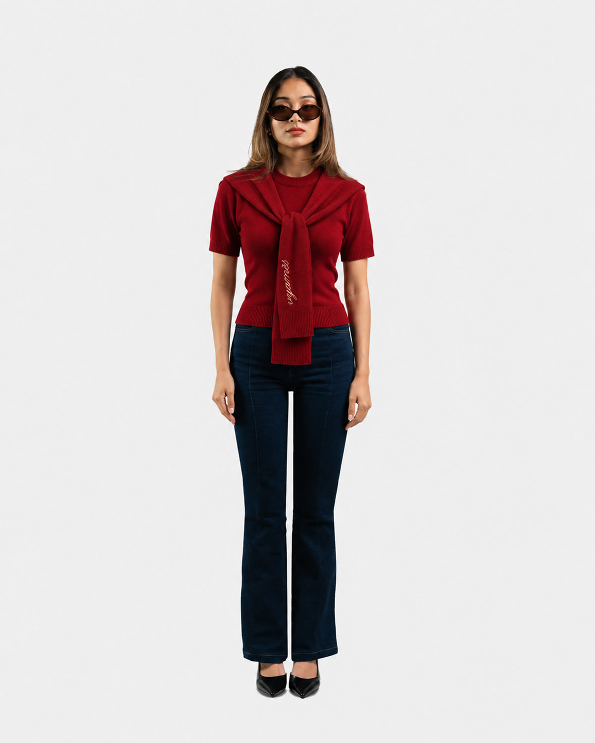 A model stands against a light gray background wearing a short-sleeve red knit top with a large draped collar detail and matching tie, paired with dark blue flared pants and black shoes.
