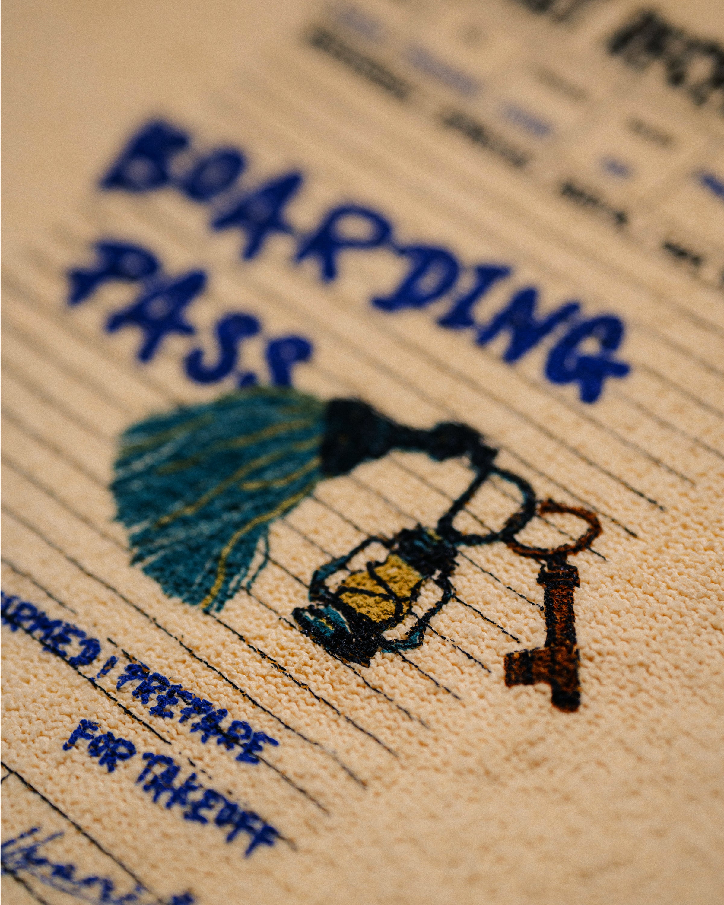 Close-up of a beige woven or embroidered surface with blue handwritten-style text reading 'BOARDING PASS,' plus a small illustrated lantern and key in dark blue, yellow, and brown.
