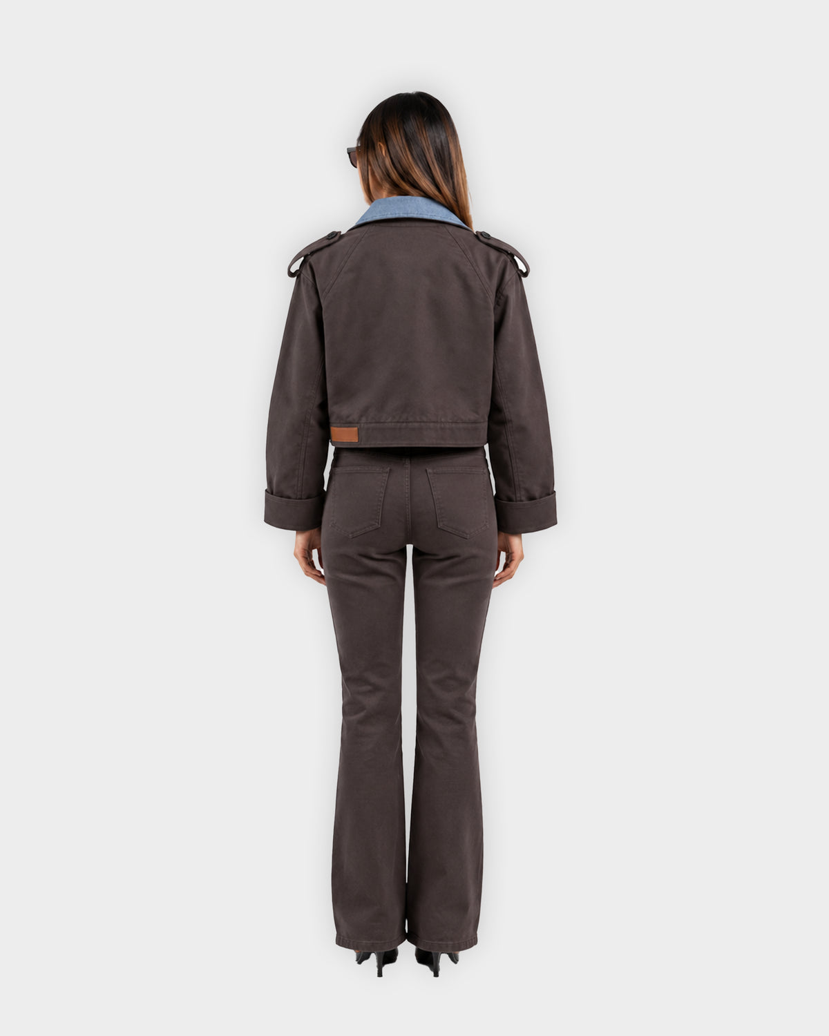 A model stands against a white background wearing a black leather moto jacket over a gray shirt, paired with dark gray patchwork jeans and brown suede boots.