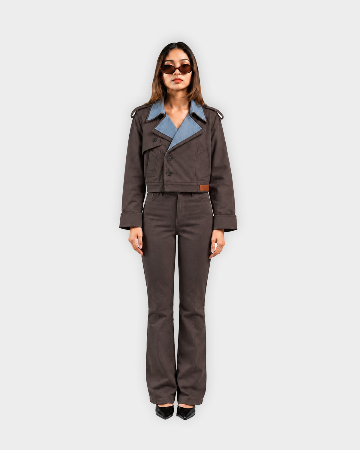 A model stands against a light gray background wearing a dark brown cropped utility jacket with a light blue collar lining and matching dark brown flared pants.